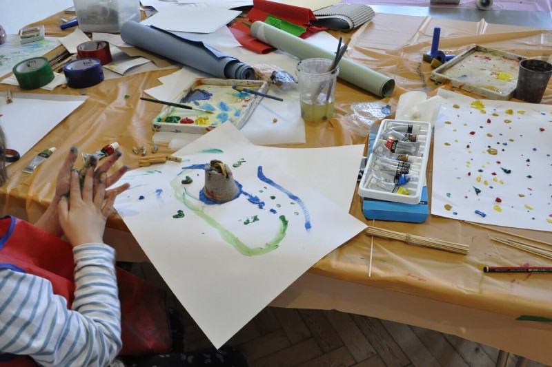 A messy arts teable with scattered paper, paints, clay shapes, splattered paint. To the left is a pair of small childrens hands wiping paint together.