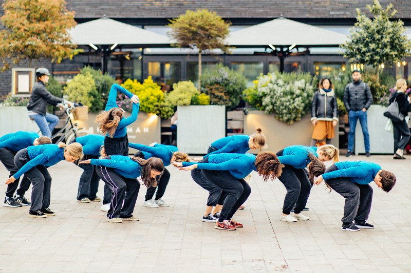 A group of dancers performing outside, all dressed in blue tops, leaning forward in a pose and one of them is standing with their arms raised