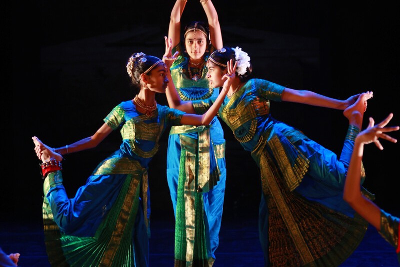 Three young women dance together on stage in Sari's. One faces the front with her hands touching above her head, the other two are facing inwards to each other