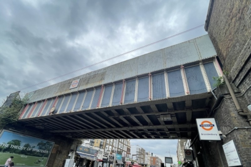 A bridge in Camden, with the london overground sign (orange) on it. The bridge is pale blue and worn out.